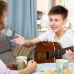 Teenager with guitar talking to mother - How to Support Your Child This School Year—Without Pushing Them to Be Perfect: Be a Gardener, Not a Carpenter - Cheerful teenage boy with guitar friendly talking to his mother at home table..