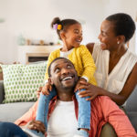 Cheerful little girl sitting on father shoulder while playing with mother at home.