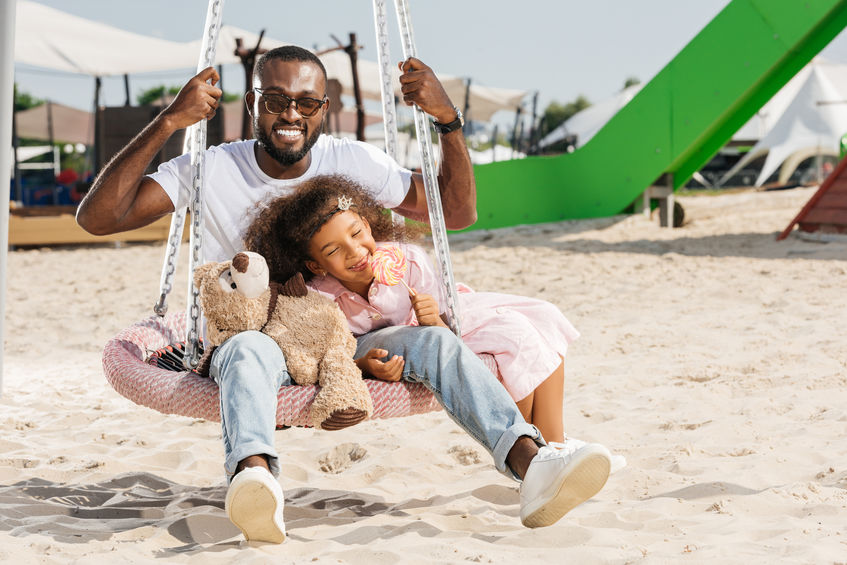 smiling african american father and daughter on spider web nest swing at amusement park